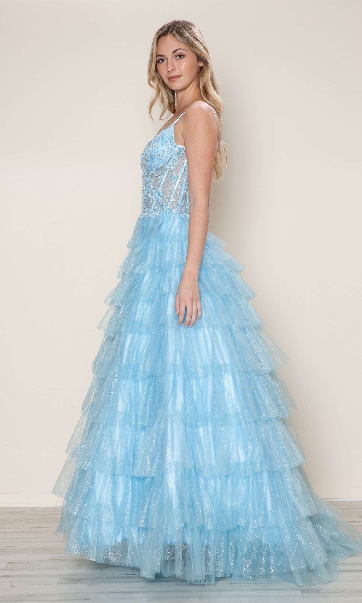 A woman stands sideways against a plain, light background wearing the Long Prom Dress: Poly USA 9608—a sleeveless, light blue gown with sparkly details, a fitted semi-sheer lace corset bodice, and a tiered tulle skirt.